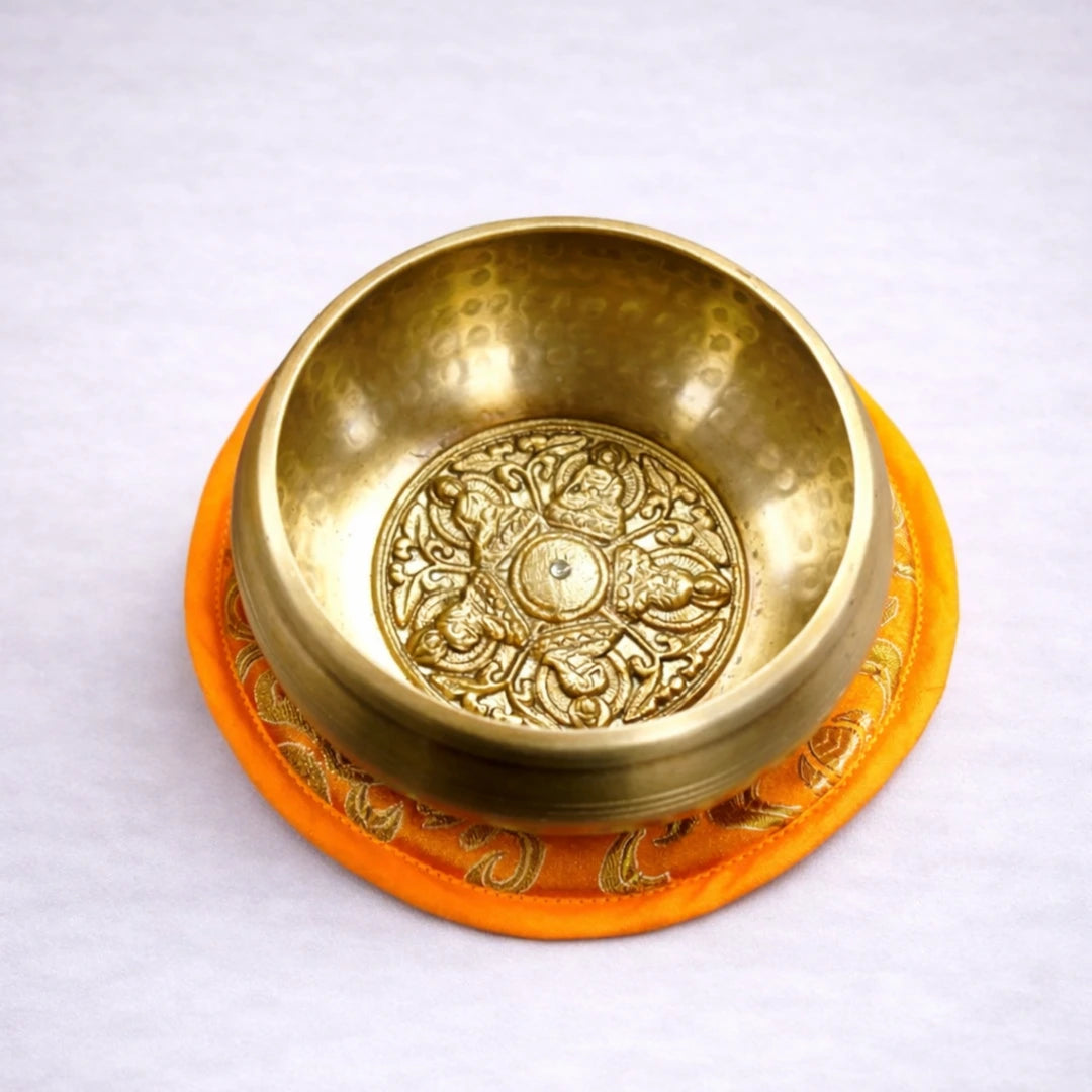Cuenco tibetano artesanal de latón con grabado interior sobre cojín naranja y fondo grisáceo suave