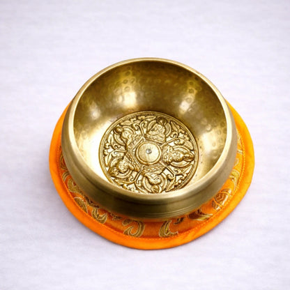 Cuenco tibetano artesanal de latón con grabado interior sobre cojín naranja y fondo grisáceo suave