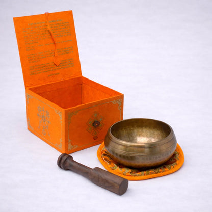 Set de cuenco tibetano artesanal con caja decorativa, mazo de madera y cojín naranja sobre fondo grisáceo suave