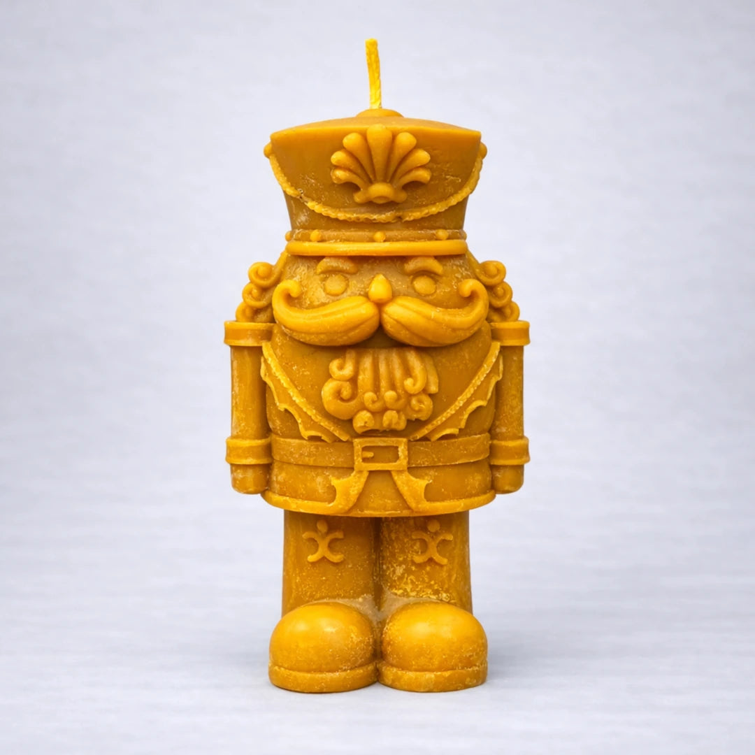 Vela artesanal de cera de abeja con forma de cascanueces decorativo, fotografiada sobre fondo gris suave, diseño detallado y acabado natural.