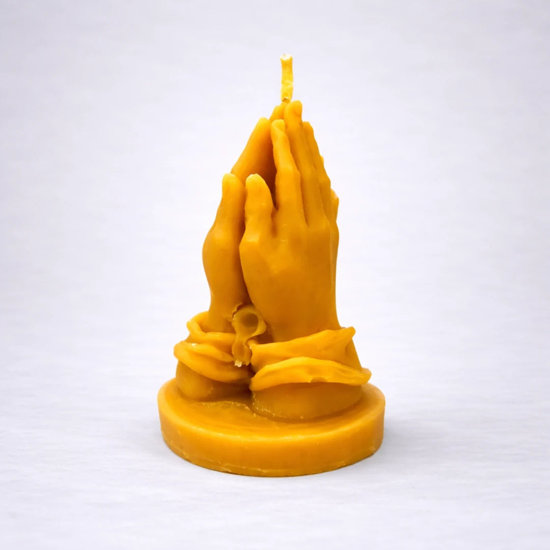 Vela artesanal de cera de abeja con manos en posición de oración, símbolo de calma, fe y conexión espiritual, sobre fondo gris suave.
