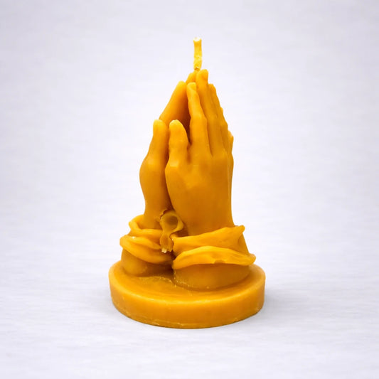 Vela artesanal de cera de abeja con manos en posición de oración, símbolo de calma, fe y conexión espiritual, sobre fondo gris suave.