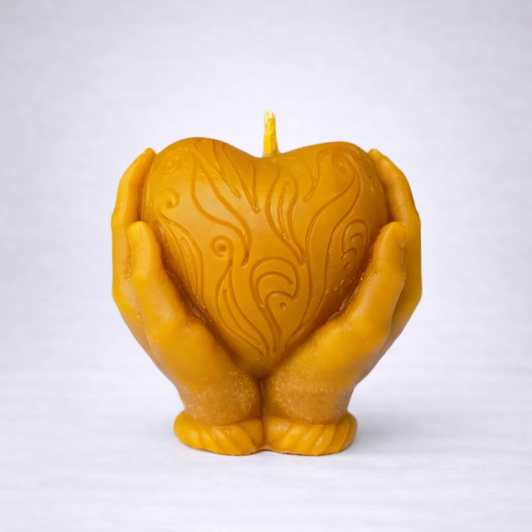 Vela artesanal de cera de abeja con manos sosteniendo un corazón, símbolo de amor, protección y energía positiva, sobre fondo gris suave.