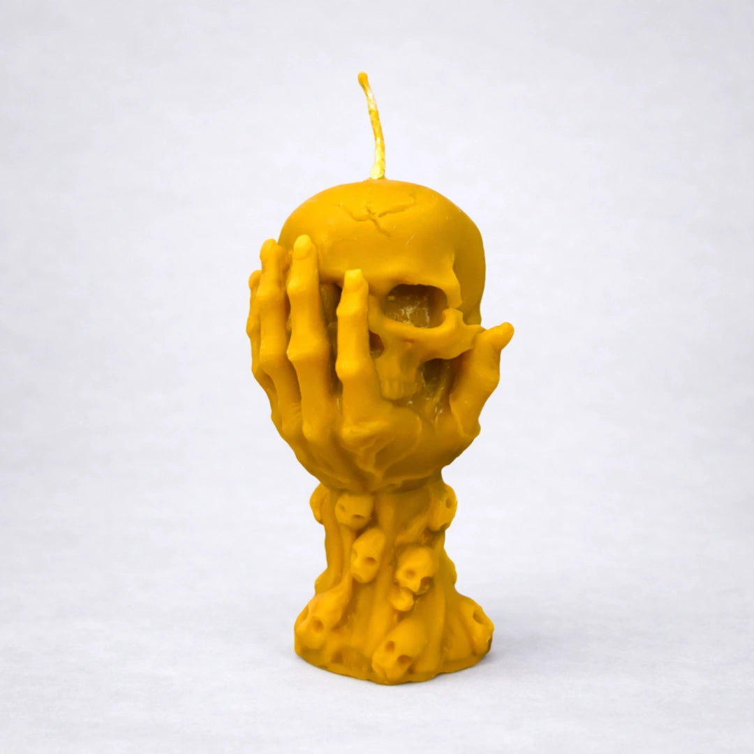 Vela artesanal de cera de abeja con diseño de calavera y manos cubriendo el rostro, símbolo de introspección y silencio interior, sobre fondo gris neutro.