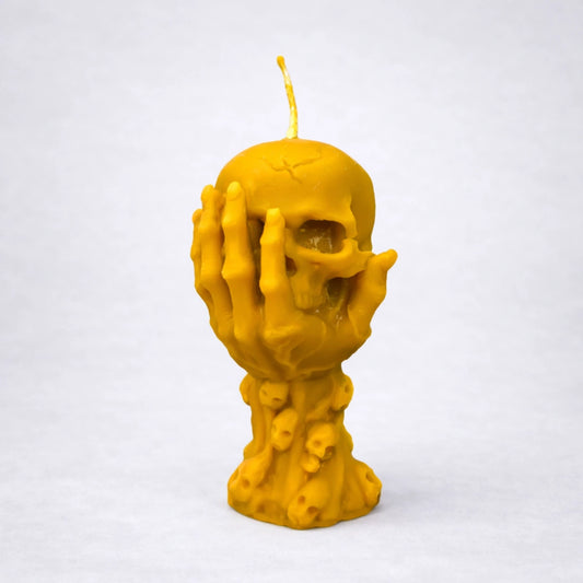 Vela artesanal de cera de abeja con diseño de calavera y manos cubriendo el rostro, símbolo de introspección y silencio interior, sobre fondo gris neutro.