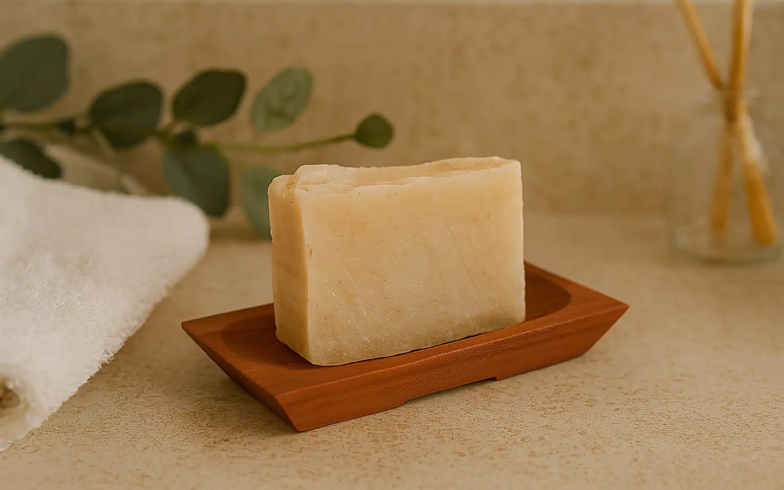 Imagen de una jabonera natural de madera con un jabón artesanal sobre una encimera de baño, acompañada de una toalla blanca, ramas verdes y difusor en el fondo, creando una escena de bienestar y cuidado personal.