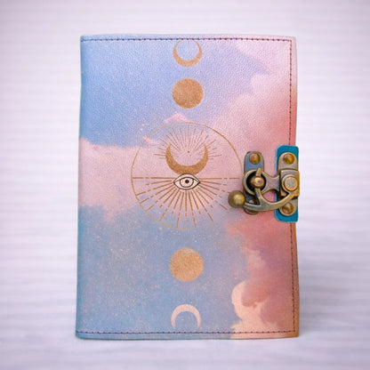 Cuaderno artesanal de cuero con diseño de fases lunares y símbolo del ojo, cierre metálico y tonos azul y rosa, ideal para journaling y escritura consciente.