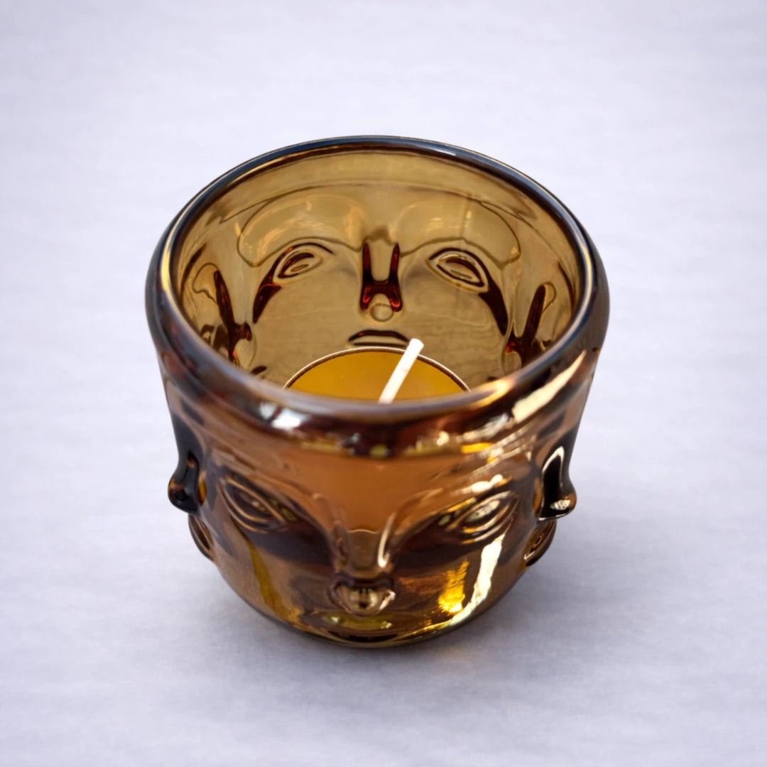 Portavelas buda de cristal ámbar con vela de té y reflejos cálidos sobre fondo gris claro